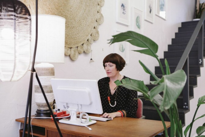 A woman with a dark bob and bangs works at a computer at a wooden desk.