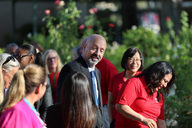 A man with medium-light skin tone and a gray and brown goatee wears a dark suit with a blue tie and smiles at the adults standing around him.