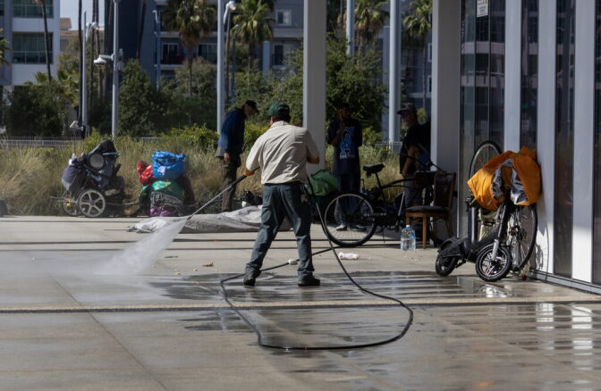 A worker sprays water on a cement floor outside a building as people with bikes and other items watch and talk among each other.