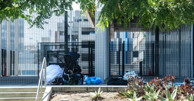 Items, including a stroller and bags, are left outside in front of a metal fence.