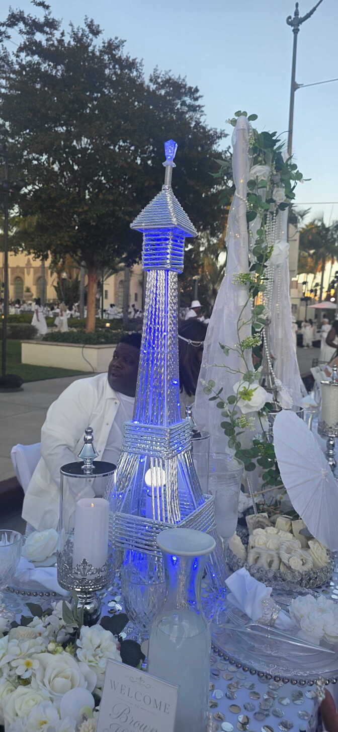 A table setting featuring a lit-up Eiffel Tower, a white candle in a silver and glass jar, a carafe of lemonade, white flowers, and a tray of white chocolate pretzels.