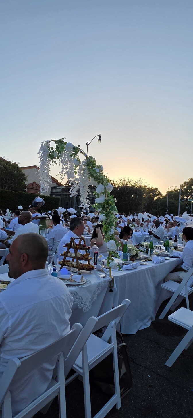 Dozens of people dressed in white sit in white folding chairs at tables with white table cloths. Glasses, plates and napkins are on the tables along with a tall flowery centerpiece.