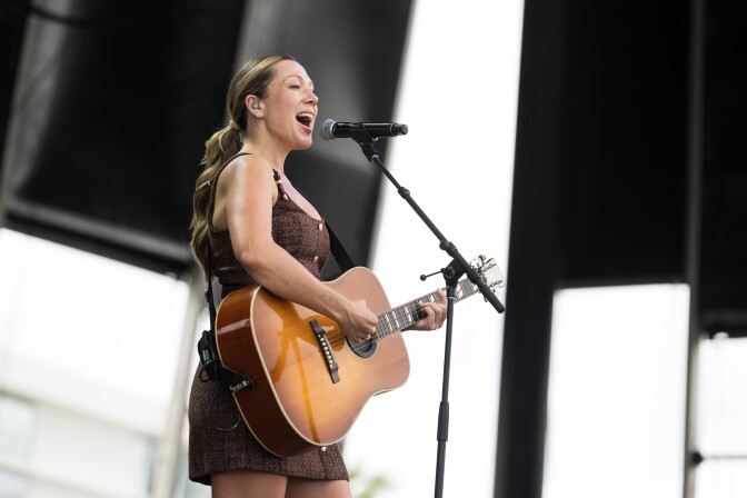 A light-skinned woman in a brown dress plays an acoustic guitar and sings onstage.