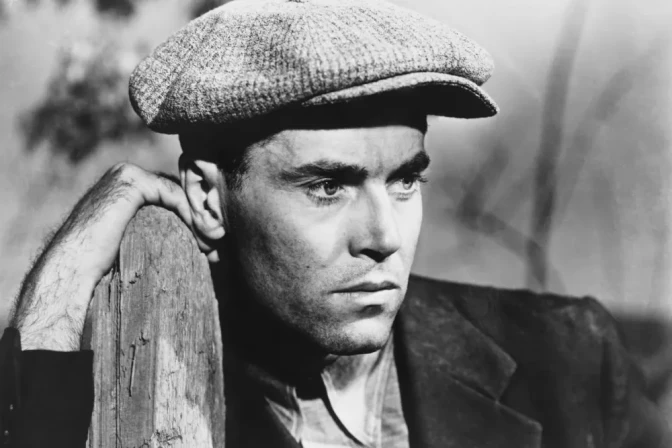 Black-and-white image of a light-skinned man wearing a hat and holding onto a fence post.