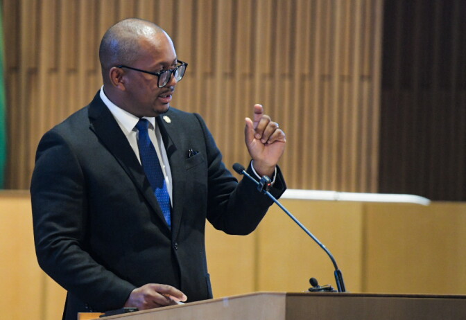Long Beach Mayor Rex Richardson, a man with medium skin tone, wearing a black suit and blue tie, speaks behind a podium as he gestures with one hand.