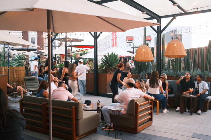 A crowd of people sit and stand on a rooftop patio surrounded by potted cacti, string lights, and umbrellas, with string lights overhead.