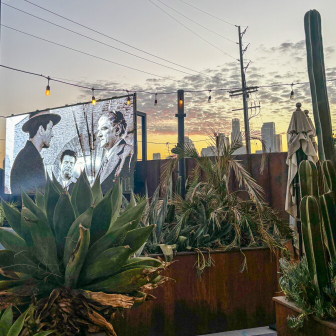 A black-and-white film plays on a large outdoor screen at dusk, framed by cacti and palm trees with the downtown Los Angeles skyline visible behind it.