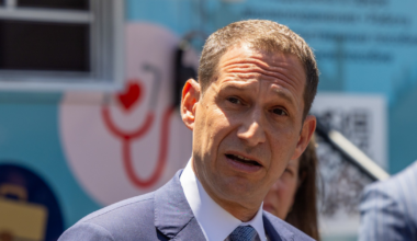 A man in a blue suit and patterned tie speaks outdoors, partially shaded, with a blurred background featuring medical-themed graphics.