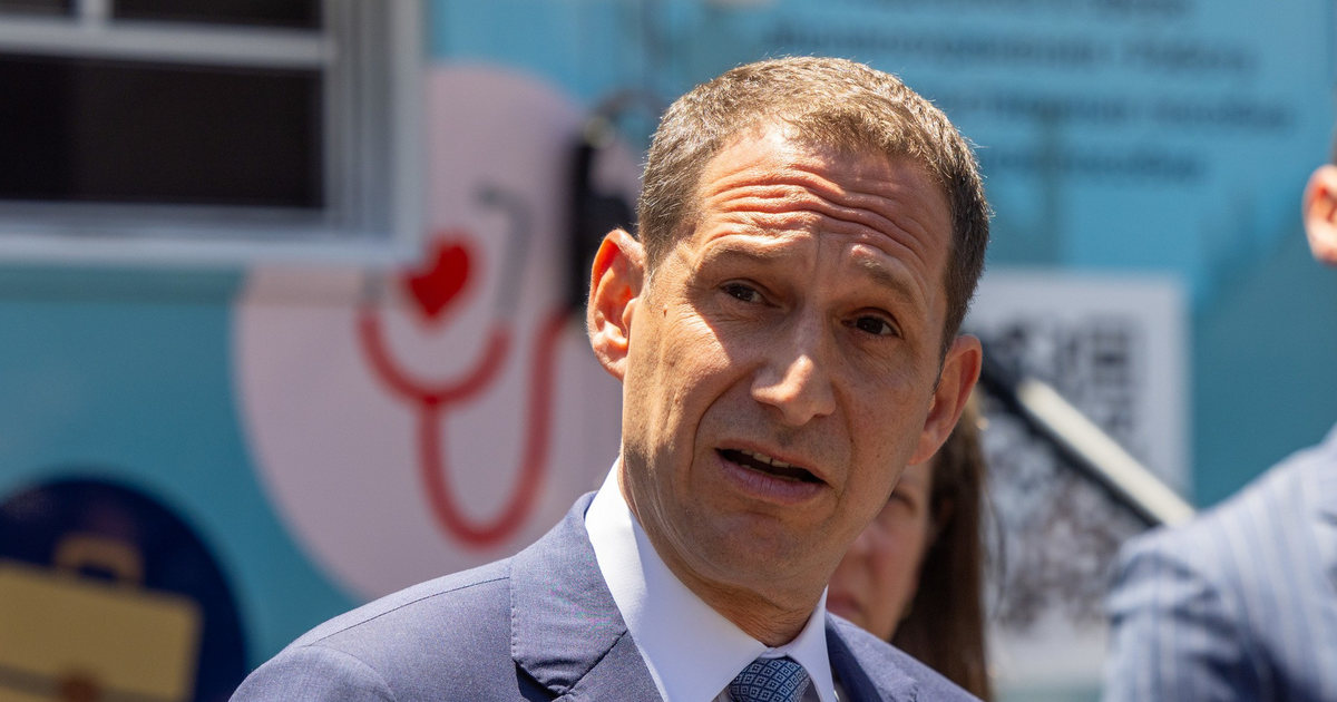 A man in a blue suit and patterned tie speaks outdoors, partially shaded, with a blurred background featuring medical-themed graphics.