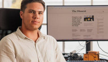 A man in a white shirt sits in an office chair with a computer monitor behind him displaying a webpage titled "The Dissent."