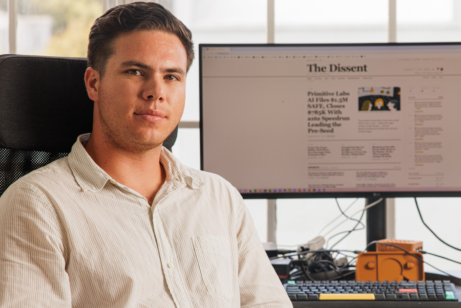 A man in a white shirt sits in an office chair with a computer monitor behind him displaying a webpage titled "The Dissent."
