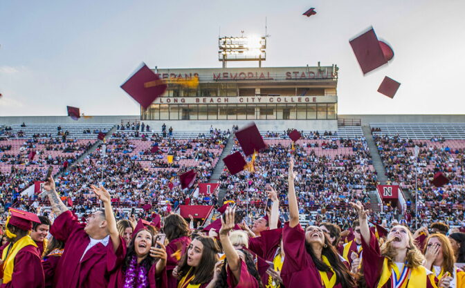 Graduates in burgundy cap and gowns through their caps in the air as people watch from stands in the background.