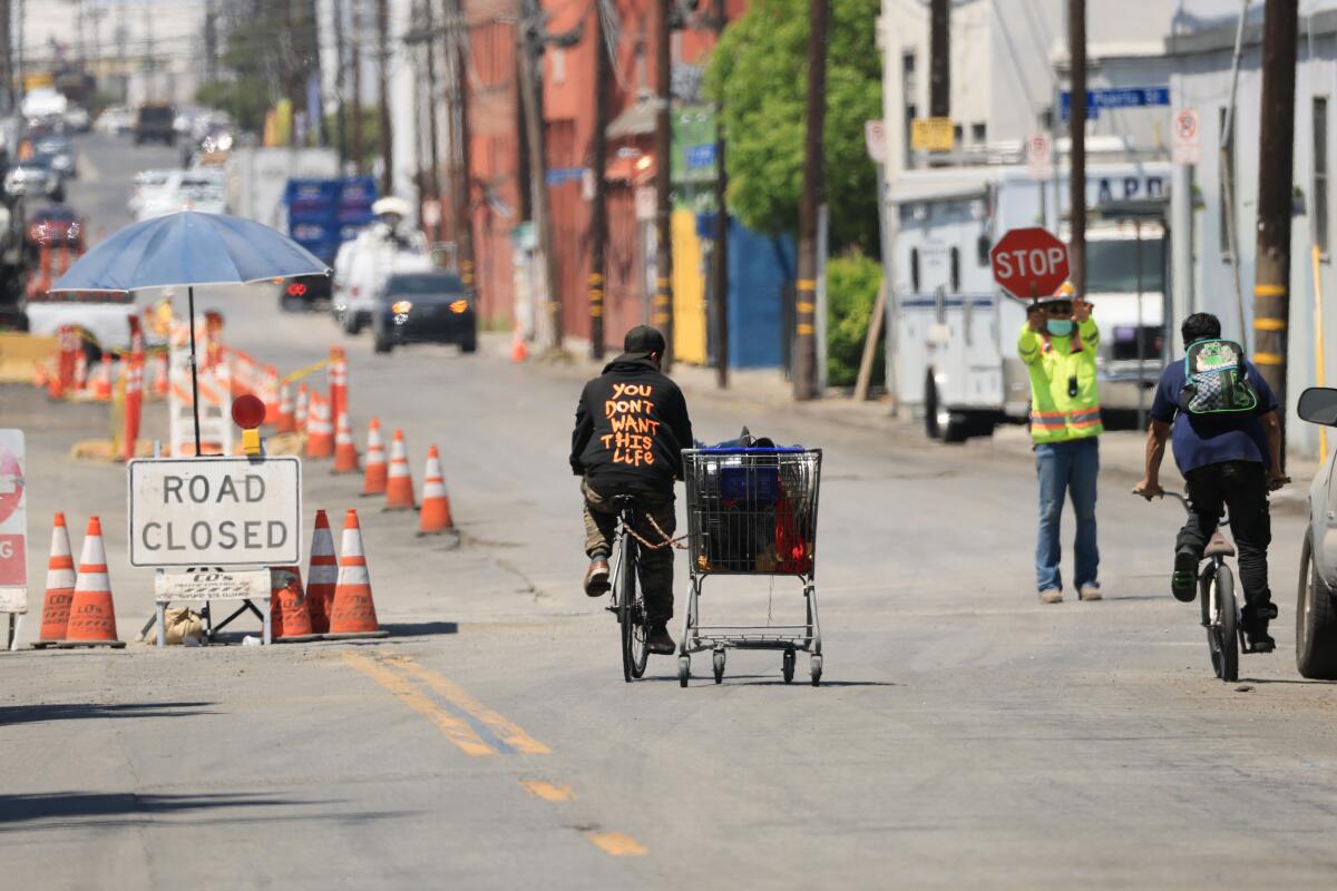 A man pulling a shopping cart full of items and a bicyclist pass construction crews