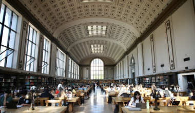 A large, ornate library reading room with high ceilings, rows of wooden tables, brass lamps, and many people studying or using laptops.