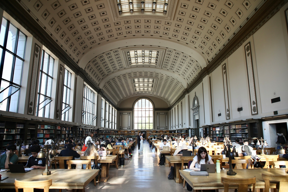 A large, ornate library reading room with high ceilings, rows of wooden tables, brass lamps, and many people studying or using laptops.