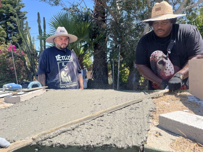 Gilbert Medrano (left) and Ryan Diaz (right) co-managers of the LA River Fingerboard Skatepark social media accounts lay down concrete on a build day. They both wear hats. Ryan wears a black graphic T-shirt, as does Gilbert. Ryan wears black gloves and various tools are seen surrounding them.