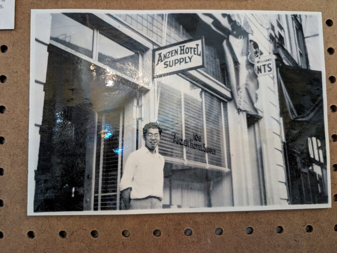 Close-up of a black and white photo affixed to a bulletin board of a young Japanese man in glasses, smiling and standing in front of a store named "Anzen Hotel Supply"