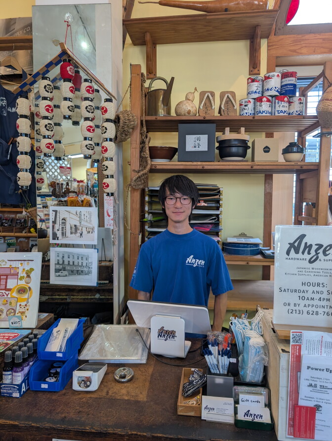 A young Japanese man in glasses and a blue shrt standing behind a cash register of a Japanese hardware store.