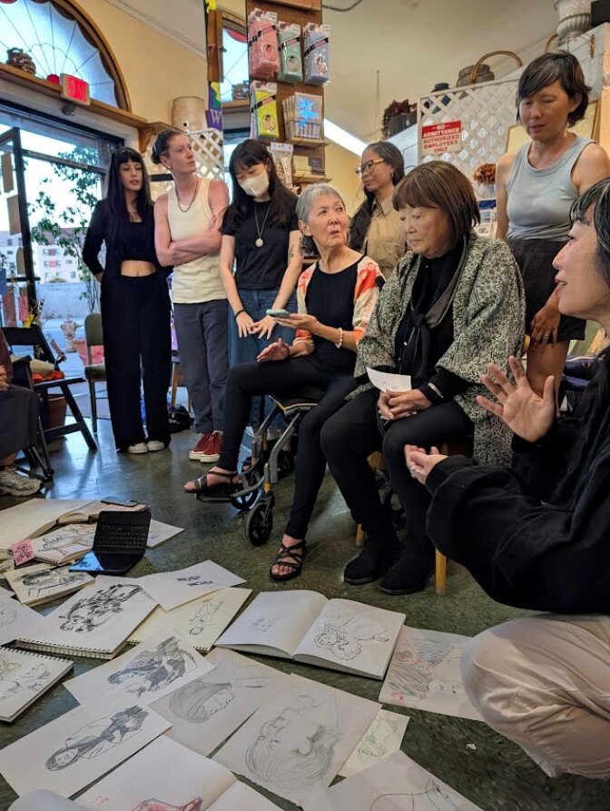 Two Japanese women in their 70s surrounded by people. They are all looking at drawings placed on the floor.