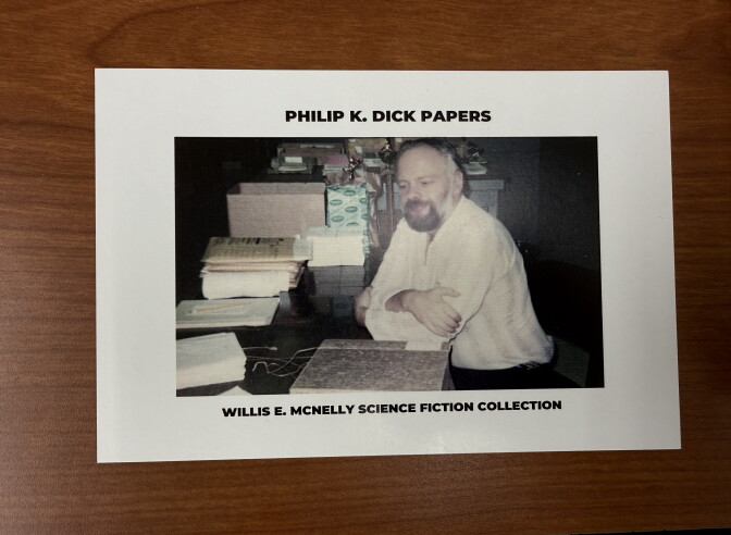 A photo depicts Philip K. Dick sitting near a stack of his literary papers.