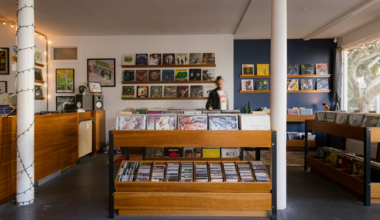 Wooden racks filled with vinyl records and CDs stand in a cozy store with framed album covers on white and dark blue walls, next to a large window.