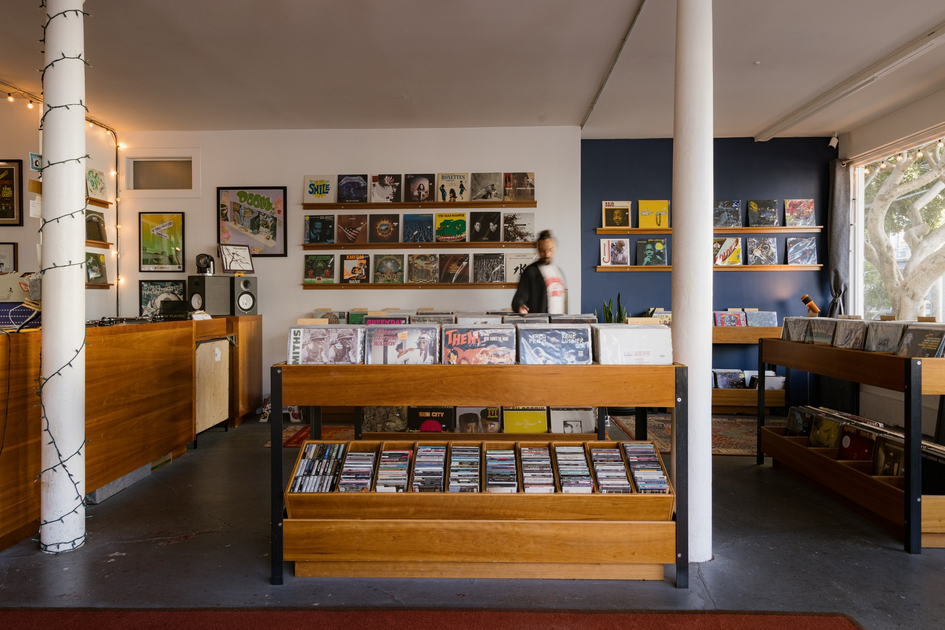Wooden racks filled with vinyl records and CDs stand in a cozy store with framed album covers on white and dark blue walls, next to a large window.