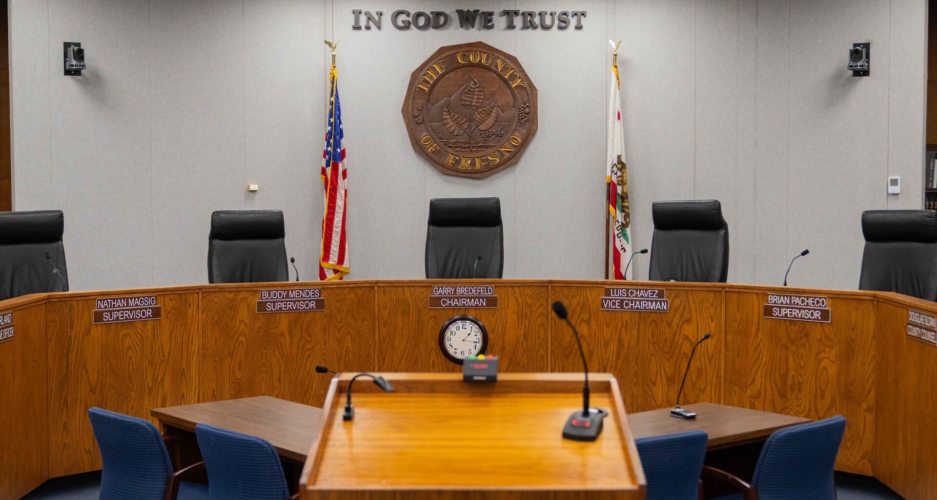 A empty government meeting room with a wooden table, chairs, and a seal on the wall.