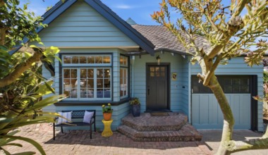 A blue house with brick steps leads to a black door; a bench with pillows and a yellow table with a plant sit beside a large window, framed by trees.