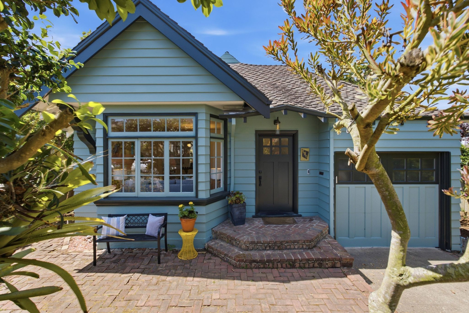 A blue house with brick steps leads to a black door; a bench with pillows and a yellow table with a plant sit beside a large window, framed by trees.