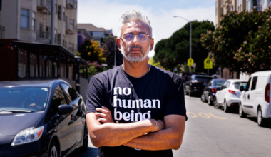 A man with silver hair and beard wears glasses and a black shirt that says "no human being," standing with arms crossed on a sunny urban street.