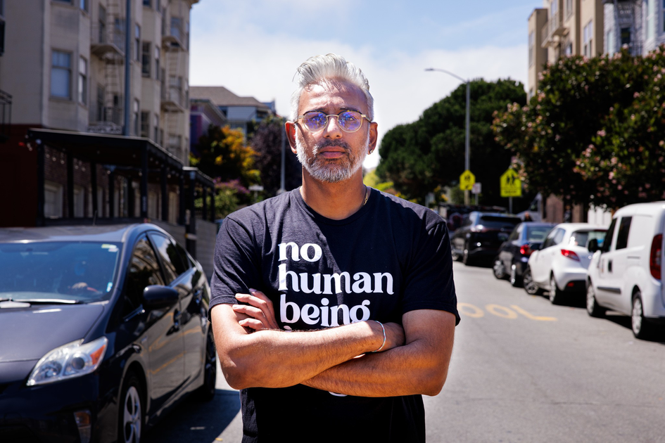 A man with silver hair and beard wears glasses and a black shirt that says "no human being," standing with arms crossed on a sunny urban street.