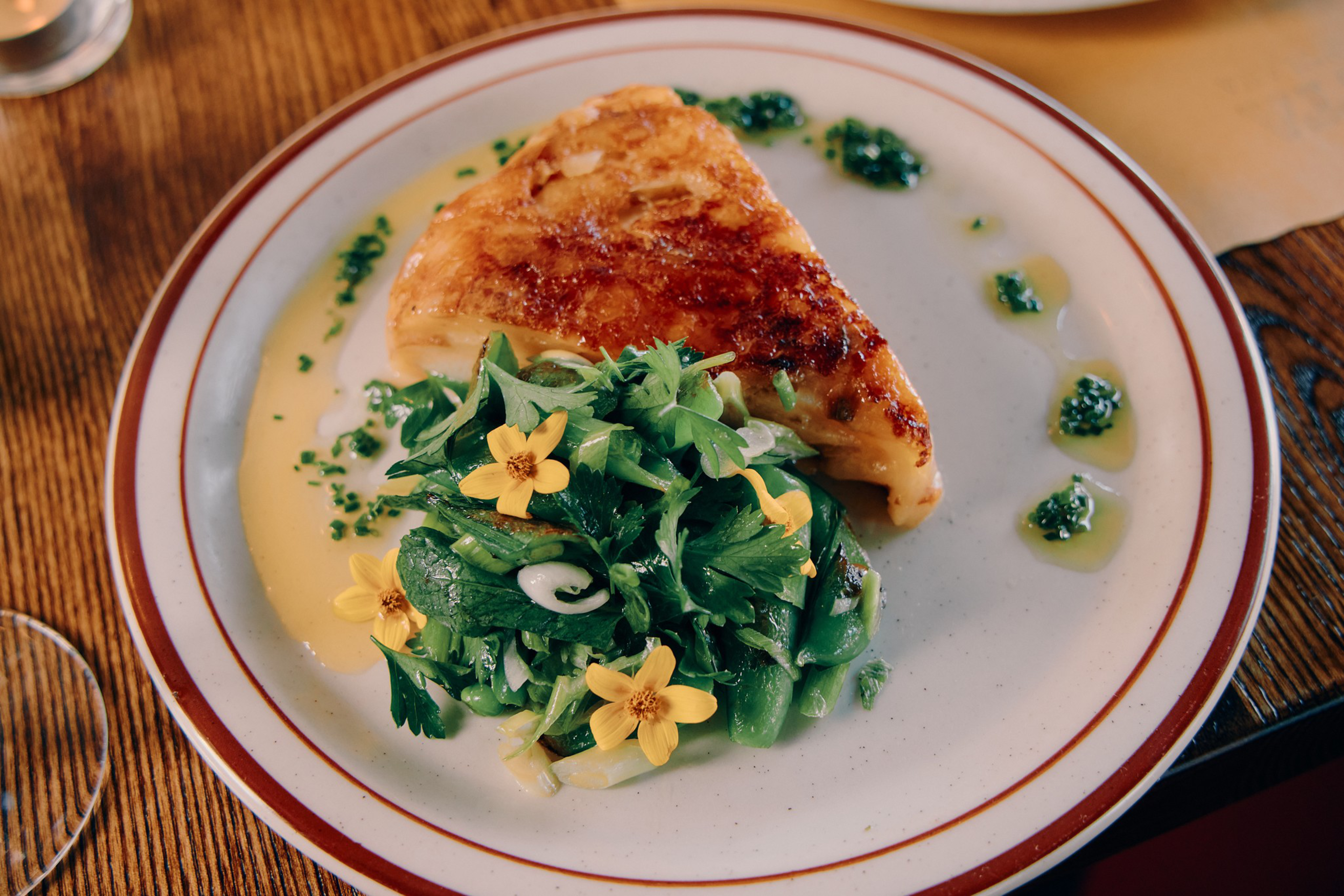 A triangular piece of grilled cheese is served with sautéed greens garnished with small yellow edible flowers and drizzled with green herb oil.
