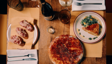 Three pieces of bread topped with meat and cheese, a round pizza, and a plate with a slice of quiche and greens are on a wooden table with drinks.