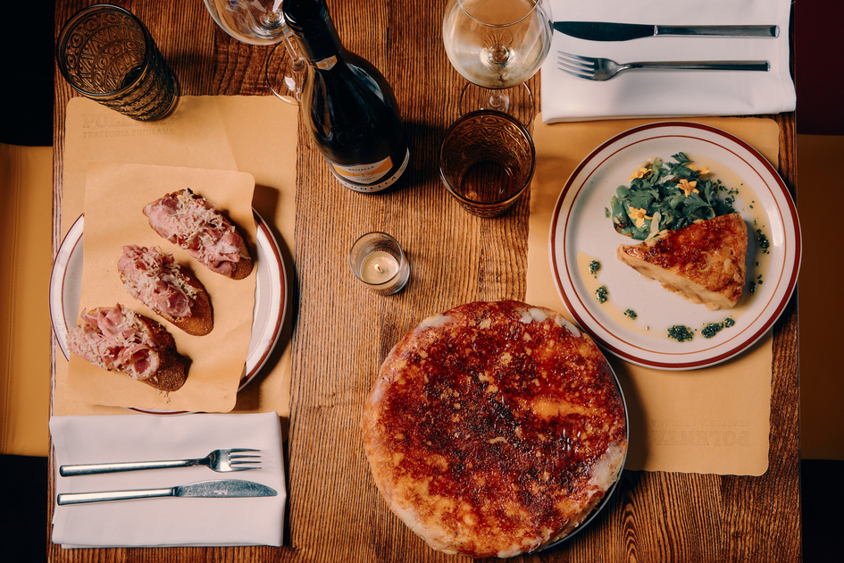 Three pieces of bread topped with meat and cheese, a round pizza, and a plate with a slice of quiche and greens are on a wooden table with drinks.