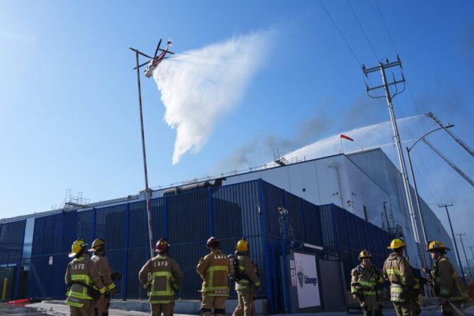 A helicopter drops water over a warehouse as firefighters watch from the ground.