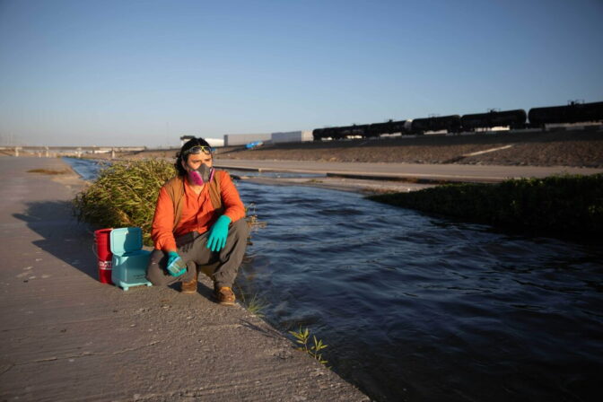 A person wearing a mask, gloves, long sleeve shirt and pants, crouches down on a cement floor next to a river with a train in the background.