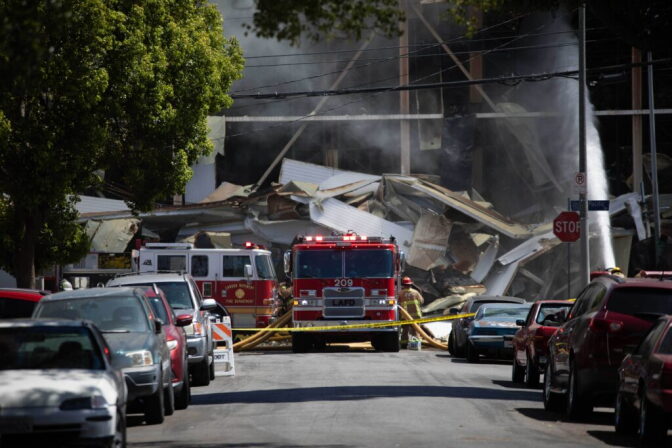 Firefighters stand next to parked firetrucks down a street in front of a warehouse on fire.
