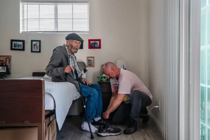 A man helps an older man sitting on a bed tie his shoes.