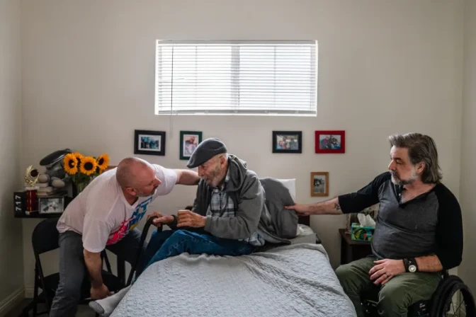 A man leaning over helps an older man sit on a bed as another mat on the other side of the twin bed helps.