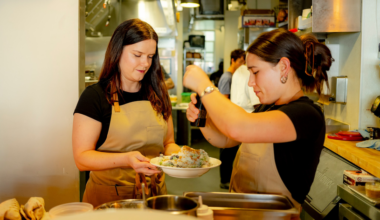 A chef wearing an apron explains a dish to diners holding wine glasses while kitchen staff prepare food in the background.