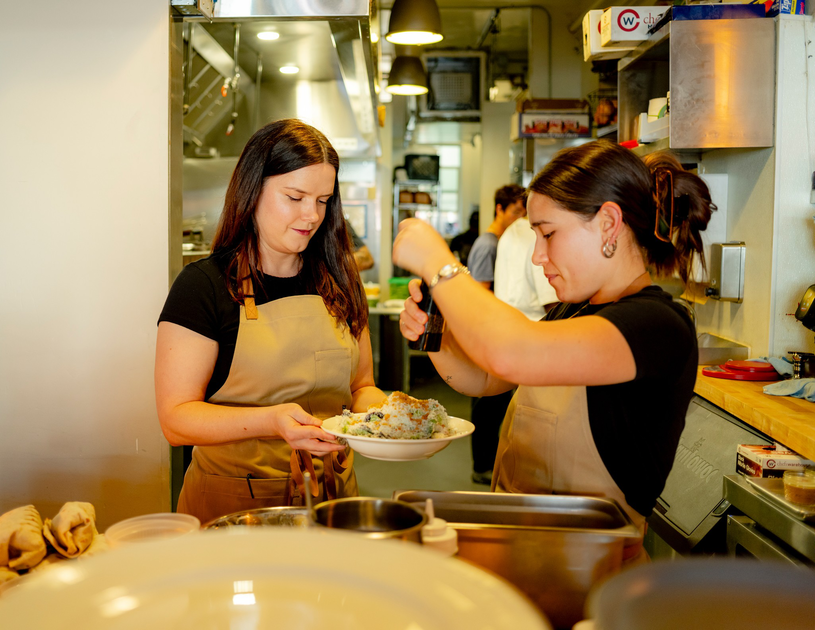 A chef wearing an apron explains a dish to diners holding wine glasses while kitchen staff prepare food in the background.