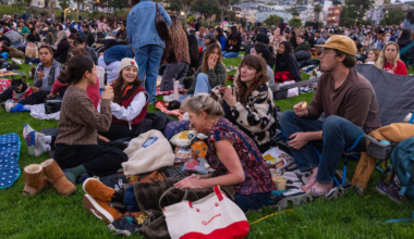 People sit on blankets and grass at a crowded outdoor event, chatting, eating snacks, and enjoying the social atmosphere.