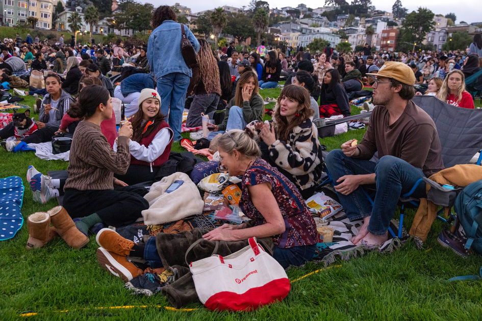 People sit on blankets and grass at a crowded outdoor event, chatting, eating snacks, and enjoying the social atmosphere.