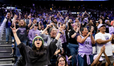 A cheering crowd at a basketball game, many wearing purple, shout and raise hands as the score shows 38 to 26 on the scoreboard.