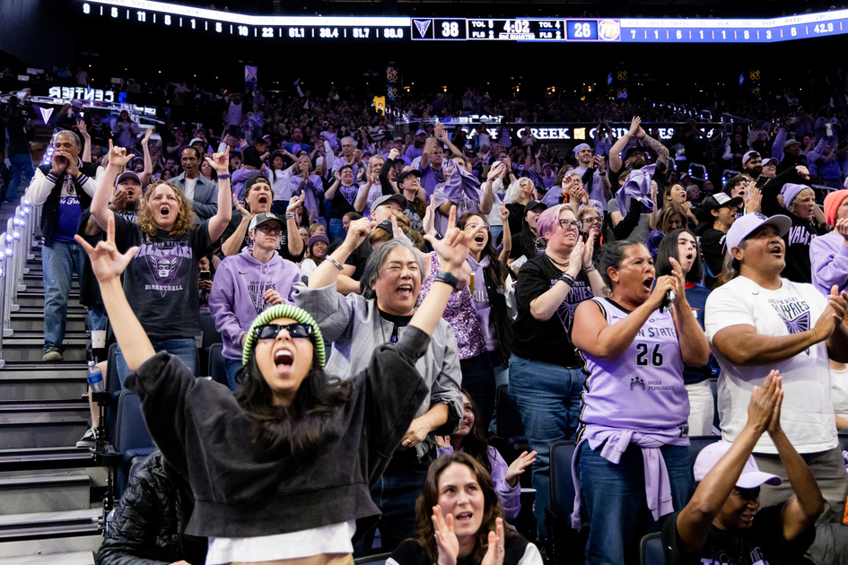 A cheering crowd at a basketball game, many wearing purple, shout and raise hands as the score shows 38 to 26 on the scoreboard.
