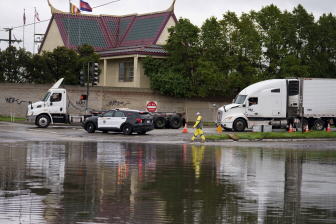 A police car blocks access to a flooded roadway as a person yellow raingear walks nearby. A semi-truck cab and semi-truck with trailer are both in the shot.