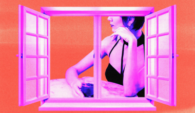 A woman in a black halter top rests her chin on one hand and holds a glass with the other, seen through an open window against an orange background.
