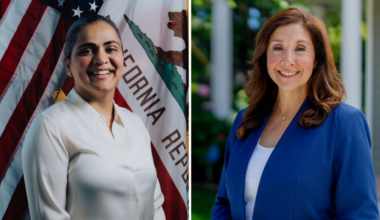 A woman in a white blouse stands smiling in front of U.S. and California flags, next to a woman with brown hair wearing a blue blazer and white top outdoors.