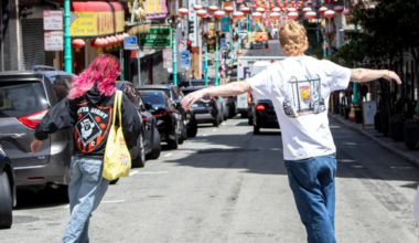 A man wearing glasses, a gray hoodie with "OUTTA SIGHT" printed on it, and navy pants stands on a sunny sidewalk holding a skateboard.
