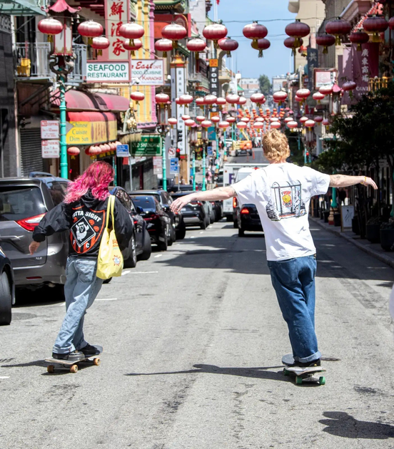 A man wearing glasses, a gray hoodie with "OUTTA SIGHT" printed on it, and navy pants stands on a sunny sidewalk holding a skateboard.
